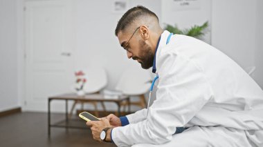 Laboratuvar önlüğü giymiş sakallı bir adam, beyaz, minimalist bir odada akıllı telefonuna gömülür..