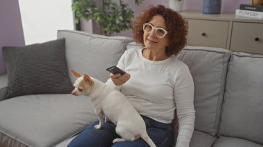 Elderly woman sitting on a couch in her living room holding a remote control with her chihuahua on her lap. clipart