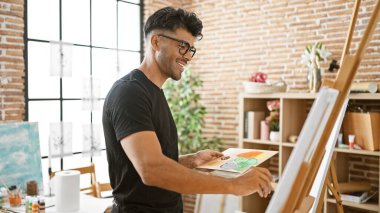 Resim stüdyosunda resim yapan yakışıklı İspanyol adam siyah gömlek ve gözlük takıyor.