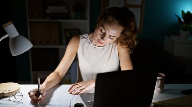İyi aydınlatılmış bir ofiste geç saatlere kadar çalışan beyaz bir kadın dizüstü bilgisayarı ve gözlüklerinin yanına notlar yazmaya odaklanmış..