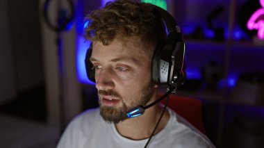 Beyaz, mavi gözlü ve sakallı genç bir adam geceleri karanlık oyun odasında kulaklık takıyor..