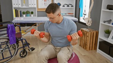 İspanyol adam rehabilitasyon kliniğinde dambıllarla egzersiz yapıyor, tekerlekli sandalyenin yanında denge topunda oturuyor..