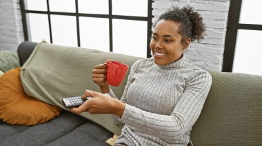 Gülümseyen bir kadın, sıcak bir oturma odasında uzaktan kumandayla kanepede kırmızı bir fincan kahveden zevk alır.
