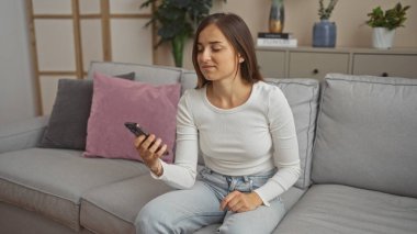 Oturma odasındaki kanepede oturan çekici genç bir kadın, rahat bir ifadeyle akıllı telefon kullanıyor..