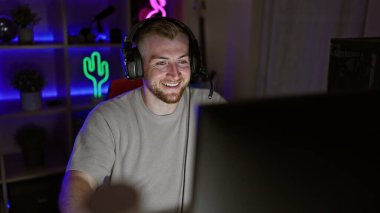 Gece vakti karanlık bir oyun odasında, sakallı, kulaklık takan, gülümseyen, beyaz bir adam..