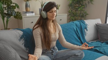 İspanyol kadın evdeki oturma odasında meditasyon yapıyor, mavi battaniyeli bir kanepede otururken kulaklık takıyor..