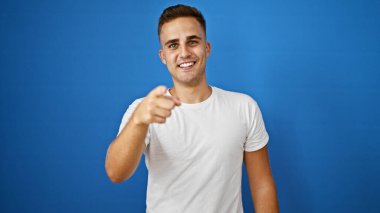 Beyaz tişörtlü, gülümseyen genç İspanyol adam, izole edilmiş mavi arka plana başparmak sallıyor..