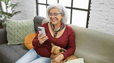 Neşeli, olgun bir kadın evcil köpeğiyle evde vakit geçirirken rahat bir kanepede akıllı telefon kullanır..