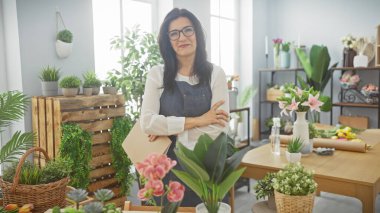 Kollarını kavuşturmuş gülümseyen orta yaşlı İspanyol kadın yemyeşil bitkiler ve renkli çiçeklerle çevrili bir çiçekçide duruyor.