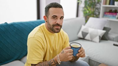 Yakışıklı, sakallı, elinde kupa tutan, evde rahat rahat oturan bir İspanyol..