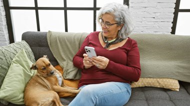 Olgun bir kadın akıllı telefon ve evcil köpekle oturma odasındaki kanepede gülümser..