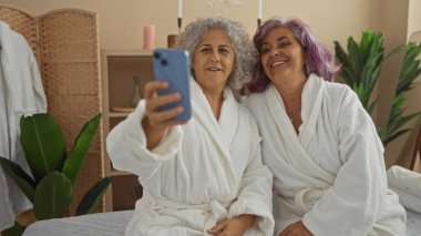Olgun kadınlar spa gününün tadını çıkarıyorlar, bornoz giyiyorlar ve selfie çekiyorlar, rahatlatıcı bir iç güzellikle çevrelenmiş.