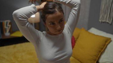 Genç, çekici, beyaz bir kadın, yatak odasında saçını bağlıyor, sarı yastıklı bir yatakta oturuyor, evde yakalandı..