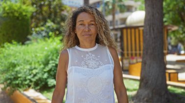 Şehir parkında açık havada duran bir kadın, güneşli bir günde yeşil ve şehir yapılarıyla çevrili beyaz bluzlu, kıvırcık saçlı orta yaşlı bir İspanyol kadını sergiliyor.