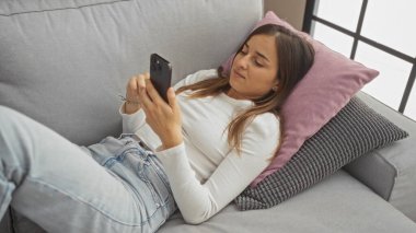 Genç ve çekici sarışın bir kadın bir apartman dairesinde kanepede uzanıyor, akıllı telefonunu kullanıyor ve yastıklara karşı dinleniyor..