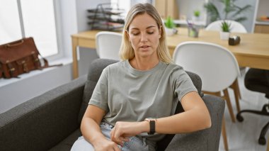 Genç sarışın bir kadın, modern bir çalışma alanında, dakiklik ve profesyonelliği çağrıştıran akıllı saatini kontrol ediyor..