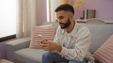 Sakallı genç İspanyol bir adam evinin rahat bir salonunda oturuyor, akıllı telefonuna odaklanmış, sıcak, davetkar bir atmosferde bir kanepe ve yumuşak yastıklarla çevrili..