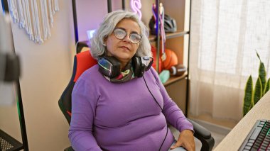 Gri saçlı, kulaklık takan olgun bir kadın iyi ışıklandırılmış bir oyun odasında oyun sandalyesinde oturuyor..