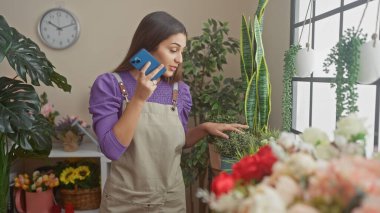 Genç bir İspanyol kadın canlı kapalı bir çiçekçide çalışırken akıllı bir telefonla konuşuyor. Etrafı taze bitkilerle çevrili..