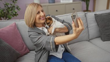 Orta yaşlı bir kadın kucağında küçük bir köpekle evde selfie çekiyor, rahat bir oturma odasında gri bir kanepede oturuyor..