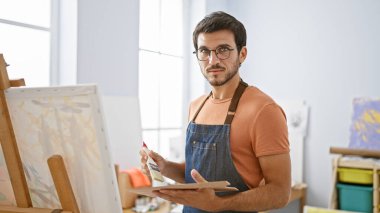 Sanat stüdyosundaki bir tuvale sakallı ve gözlüklü genç bir İspanyol adam yaratıcılık ve konsantrasyon aktarıyor..