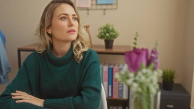 Düşünceli sarışın bir kadın yeşil kazaklı rahat bir oturma odasında kitaplar ve bitkilerle