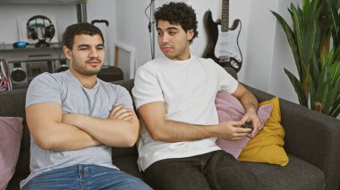 Modern bir salonda, gitarlar ve bitkilerle bir kanepede uzanmış, rahat bir atmosferi canlandıran iki adam..