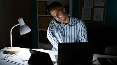 Handsome concentrated young man working late in the office using a laptop with desk lamp on, papers scattered and a tablet nearby. clipart