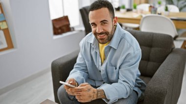 Modern bir ofis ortamında sakallı, akıllı bir telefon tutan gülümseyen İspanyol bir adam, uysal bir profesyonel ortamı canlandırıyor..