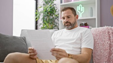 Middle-aged hispanic man with grey hair reading a paper in a modern living room, sitting on a grey sofa. clipart