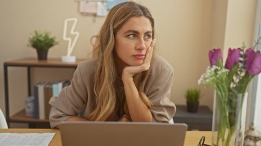 Evde dizüstü bilgisayarda çalışan, çiçekler ve dekorlarla çevrili düşünceli genç bir kadın..
