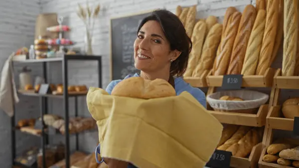 stock image Woman smiling holding fresh bread in a bakery filled with various baked goods and baguettes displayed on shelves in the background