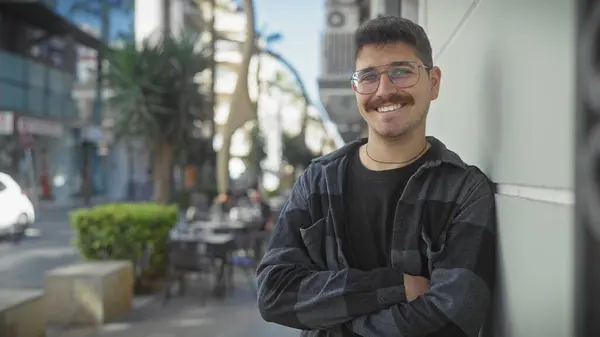 stock image A cheerful young hispanic man with a moustache standing with crossed arms on a city street.