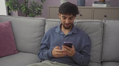 Genç, mutlu Arap adam gülümsüyor, evdeki koltukta oturuyor, akıllı telefon kullanırken el ve parmaklarıyla işaret ediyor.