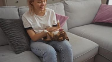 Genç sarışın bir kadın, sıcak bir arkadaşlık ve rahatlık anı sergileyen, gri bir salonda, kanepede Chihuahua köpeğiyle oynarken gülümsüyor..