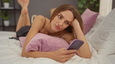 Genç sarışın bir kadın, yatak odasındaki bir yatakta uzanıyor. Akıllı telefon kullanırken hayal kırıklığına uğramış görünüyor. Yastıklarla çevrili, modern bir iç mekan..