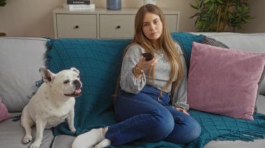 Genç, güzel, sarışın bir kadın evcil köpeğiyle bir kanepede oturuyor. Uzaktan kumandayla ev ortamını yakalıyor..