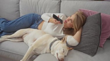 Genç bir kadın evcil köpeğiyle bir kanepede uzanıyor, elinde akıllı bir telefon tutuyor ve sıcak bir oturma odasında mesaj kaydediyor..