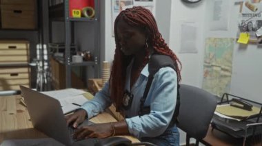 Bir polis dedektifinin ofisinde dizüstü bilgisayarda çalışan odaklanmış Afro-Amerikalı kadın. Etrafı dosyalar ve ekipmanlarla çevrili..
