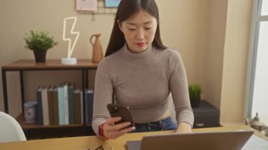 Genç Asyalı bir kadın modern bir apartman dairesinde dizüstü bilgisayarı ve dekoratif bitkileri olan bir akıllı telefon kullanıyor..