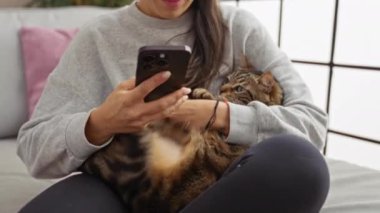 Oturma odasındaki kanepede oturan bir kadın akıllı bir telefon tutarken neşeli bir tekir kedi kucağına sarılıp rahat, sade bir atmosfer yaratıyor..