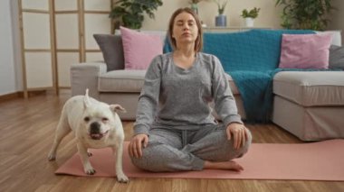 Sarışın genç bir kadın oturma odasında köpek evcil hayvanıyla meditasyon yapıyor. Etrafı rahat mobilyalar ve doğal aydınlatmalarla çevrili..