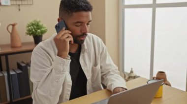 Modern evde dizüstü bilgisayarla çalışırken akıllı telefon kullanan odaklanmış genç bir adam..
