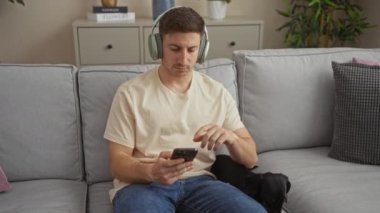 Kulaklık takan genç İspanyol bir adam oturma odasında bir daksund ile oturuyor, akıllı telefonunu kullanıyor..