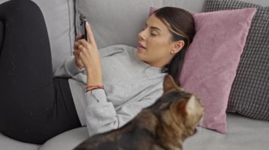 Genç bir İspanyol kadın, evcil kedisi yakınlardaki bir oturma odasında otururken bir koltuğa uzanıp telefonunu kullanıyor..