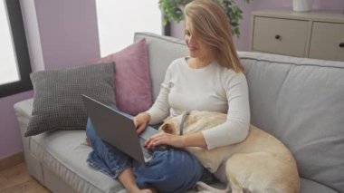 Mutlu bir kadın dizüstü bilgisayarıyla kutlama yapıyor ve evinde rahat bir kanepede bir köpek var.