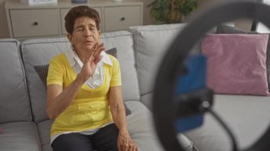 Rahat bir oturma odasındaki kanepede oturan yaşlı bir İspanyol kadın, halka ışığına monte edilmiş akıllı bir telefonla iletişim kuruyor ve içten bir gülümsemeyle el sallıyor..