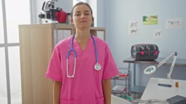 Pembe önlüklü kadın veteriner boynunda steteskopla klinik odasında güvenle duruyor. Arka planda tıbbi ekipmanlar ve hayvan posterleri var..