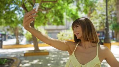 Parlak şehir parkında yeşil ağaçlarla selfie çeken, sarı bluzlu, güneşli şehirde açık havada dururken gülümseyen, kısa esmer saçlı ve güzel bir kadın.