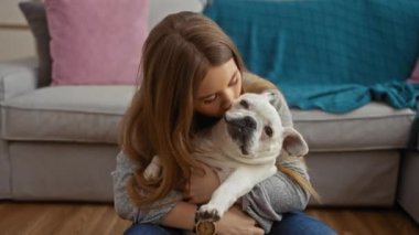 Sarışın genç bir kadın, rahat bir oturma odasında oturmuş, rahat bir ev dekoruyla çevrili olan bağlarını ve sevgilerini vurgularken beyaz köpeğini öpüyor..